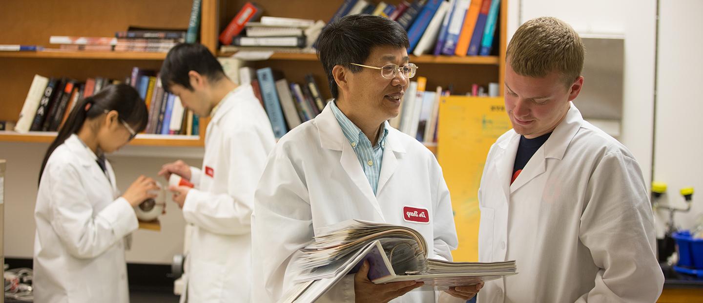 four people in white lab coats, standing in front of a book shelf, looking through a binder