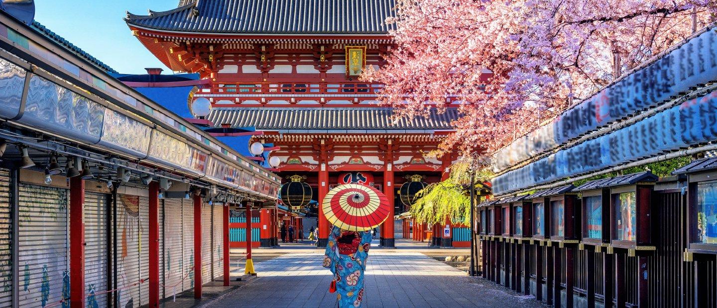 在日本，一个人撑着红白相间的伞，走在建筑物之间的小路上.