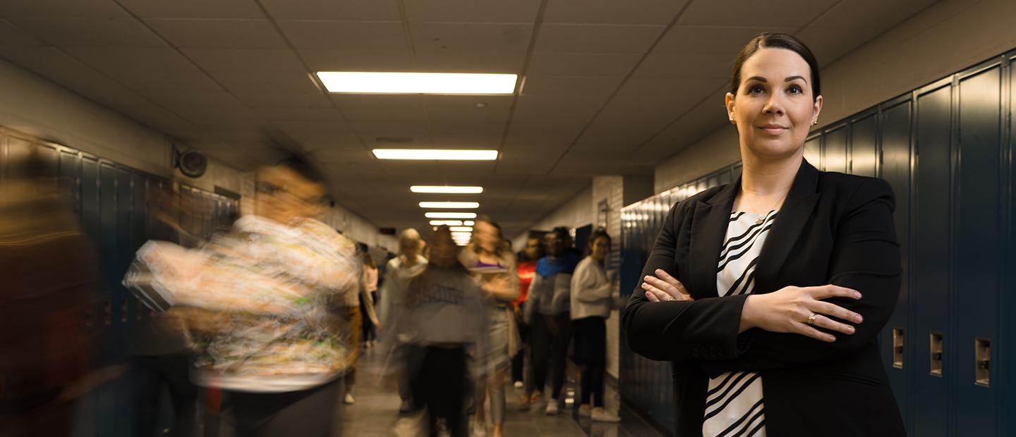 一个女人站着，双臂交叉在胸前, 在一所学校的走廊里，蓝色的储物柜沿着墙壁排列，人们跟在她后面.