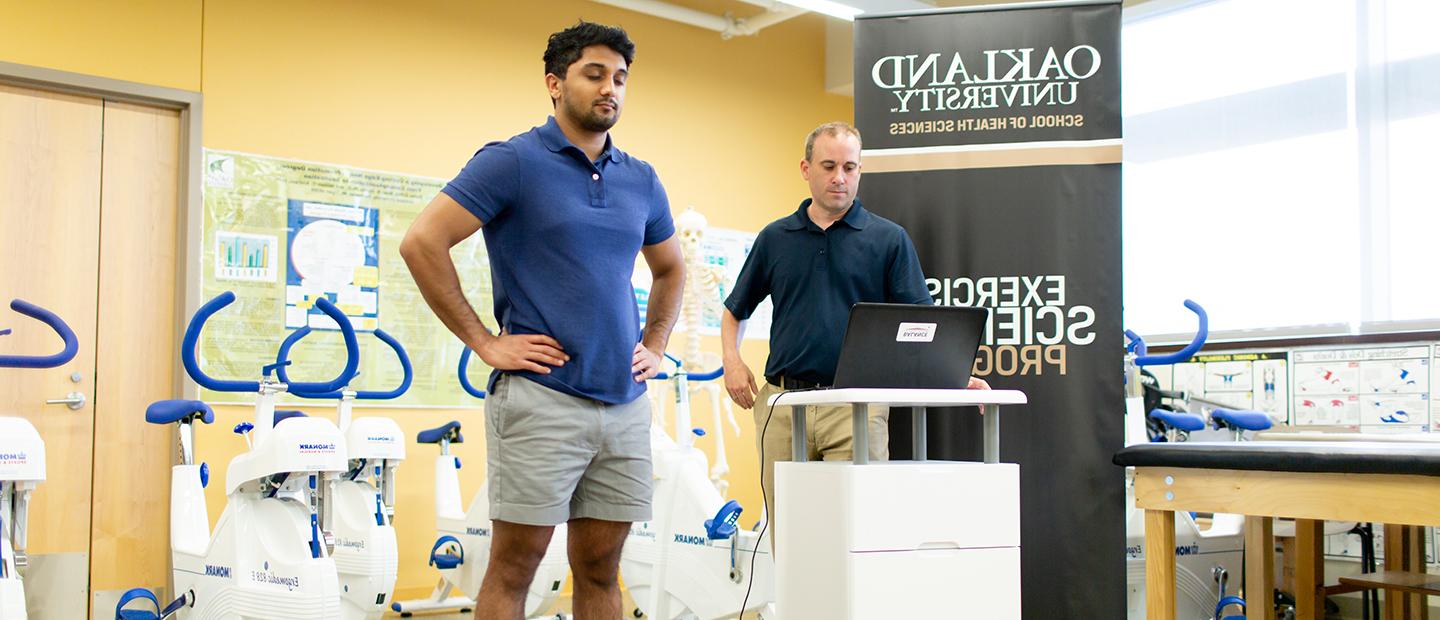 在健身房里，一名男子站在称上，另一名男子在电脑屏幕上读取数据.
