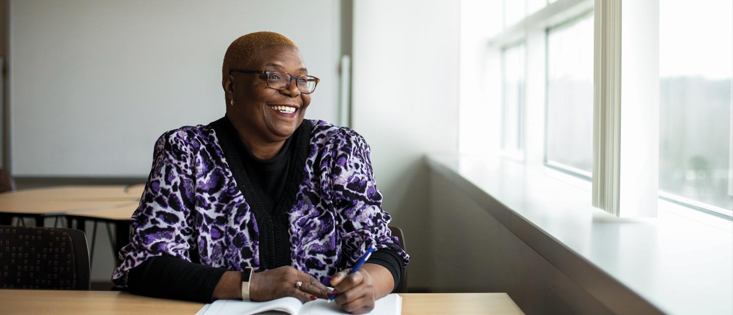 一位非洲裔美国妇女坐在办公桌前，对着她旁边的窗户微笑